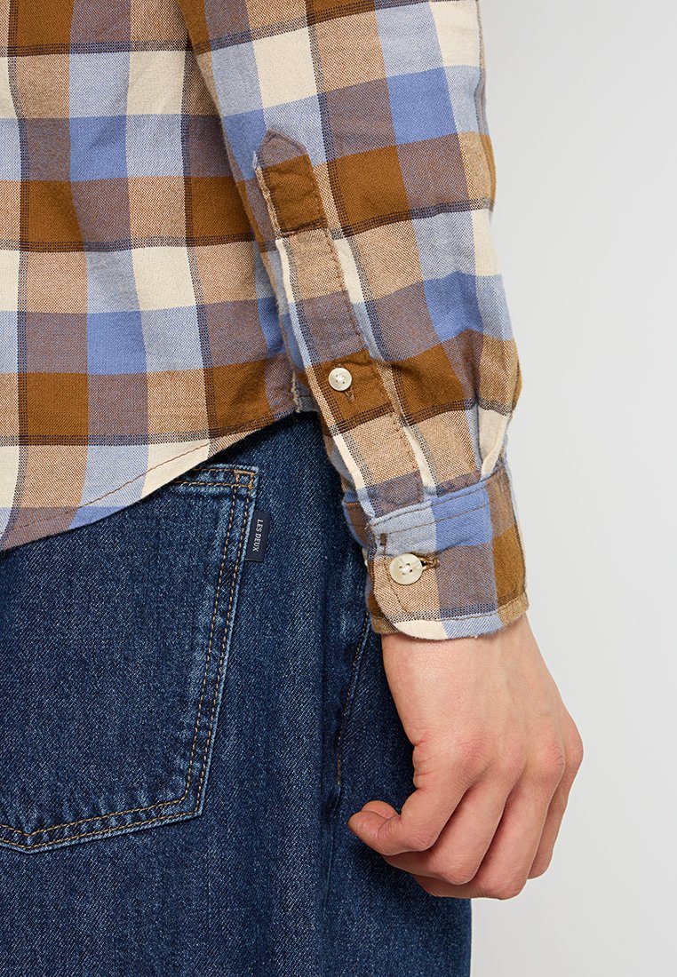 Camicia a quadri marrone, blu e crema con bottoni sui polsini, realizzata in un tessuto morbido, abbinata a jeans in denim scuro e con una tasca visibile.