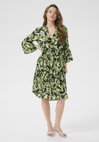 Grünes und schwarzes Blumenkleid mit V-Ausschnitt, langen Glockenärmeln und gestuftem Design, aus leichtem Material hergestellt. Tragen mit weißen Absatzsandalen.