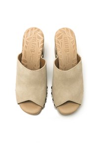 Mules abiertos de ante beige, sin tacón, con suela de goma, que presentan una plantilla de cuero marrón texturizada y un diseño ondulado en el borde superior.