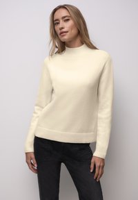 Žena se světle hnědými vlasy, oblečená v krémovém svetru a černých kalhotách, stojící před jednoduchým světle šedým pozadím.