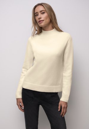 Kobieta o jasnobrązowych włosach, ubrana w kremowy sweter i czarne spodnie, stojąca na tle jednolitego jasnoszarego tła.