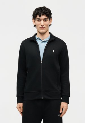 Jonge man met kort krullend haar, gekleed in een zwarte jas met rits over een lichtblauw overhemd met kraag, staand tegen een effen achtergrond.