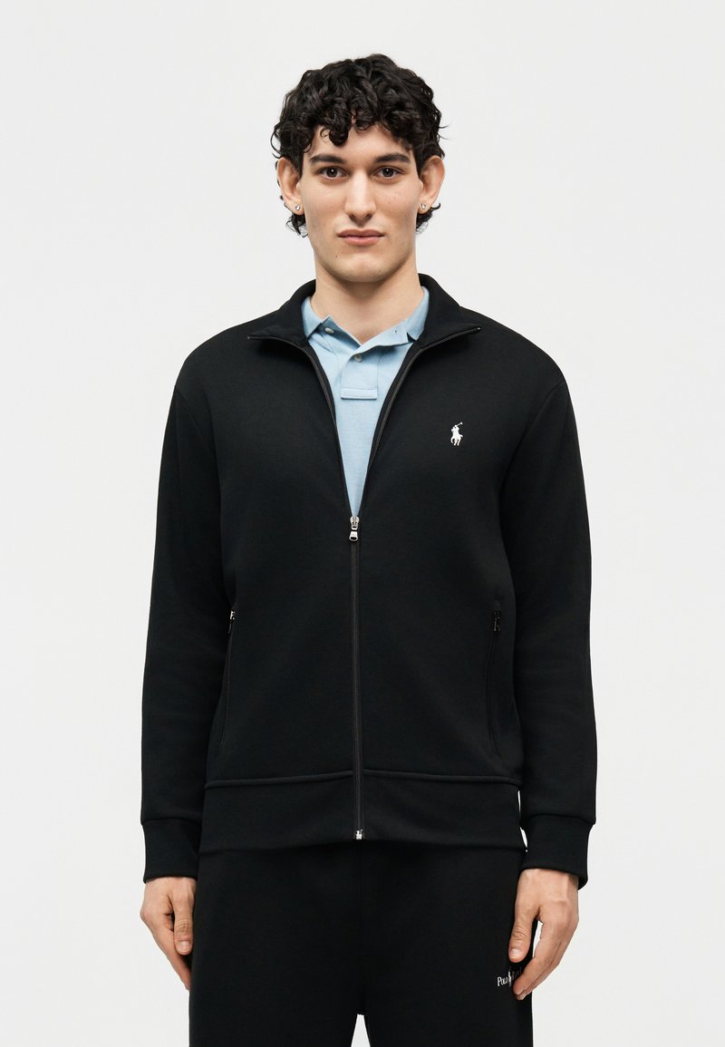 Mladý muž s krátkými kudrnatými vlasy, který má na sobě černou zipovou bundu přes světle modrou košili s límečkem, stojící před jednobarevným pozadím.