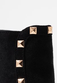 Bottine en daim noir ornée de clous pyramides dorés sur le côté, avec un bord supérieur arrondi et une texture lisse.