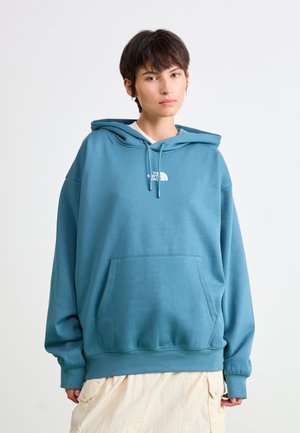 Sweatshirt à capuche bleu en tissu doux, avec une poche avant, des poignets côtelés et un petit logo blanc sur la poitrine.
