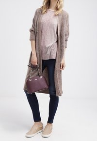 Cardigan lungo di color nocciola chiaro sopra un top rosa con trama, jeans scuri e scarpe slip-on beige. Tiene in mano una piccola borsa a tracolla di color viola scuro con dettagli in catena.