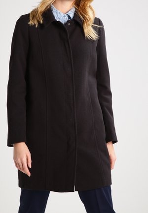 Femme portant un manteau noir longueur genou sur une chemise bleu clair à motifs et un pantalon foncé, debout sur un fond uni.