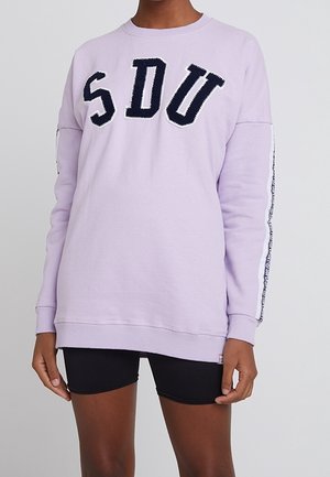Person iført en lyse lilla sweatshirt med bogstaverne "SDU" foran og sorte shorts mod en ensfarvet baggrund.