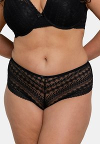 Sous-vêtements en dentelle noire avec un motif géométrique, présentant des panneaux transparents, des bords festonnés et un nœud décoratif à l'avant.