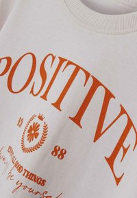 Weißes Sweatshirt mit fettgedrucktem orangefarbenem Text "POSITIVE" und floralen grafischen Details. Rippkragen und glatte Baumwolltextur.