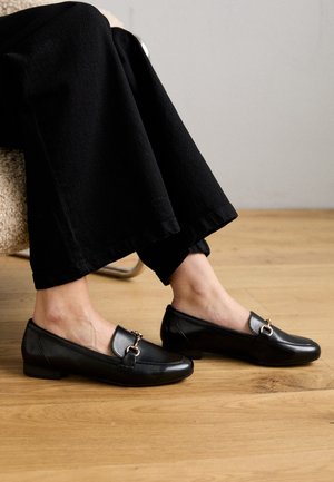 Sorte læderloafers med lav hæl, der har en guldfarvet metaldekoration foran, stylet med sorte bukser med vide ben.