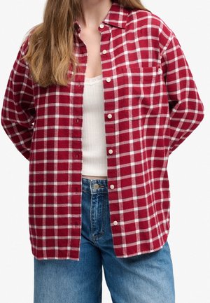 Femme portant une chemise à carreaux rouge et blanche déboutonnée par-dessus un débardeur blanc et un jean bleu sur un fond blanc uni.