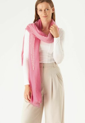 Écharpe rose en matériau doux avec des bords effilochés, drapée de manière décontractée sur un haut blanc, mettant en valeur une texture légère et aérée.