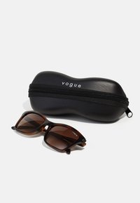 Ochelari de soare cu model țestoasă, cu lentile maronii degradate, alături de un toc oval dur, negru, cu fermoar și logo-ul alb "vogue".