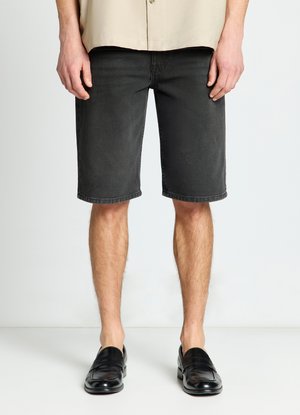 Uomo che indossa pantaloncini in denim neri, mocassini in pelle neri e una camicia beige, in piedi davanti a uno sfondo chiaro e semplice.