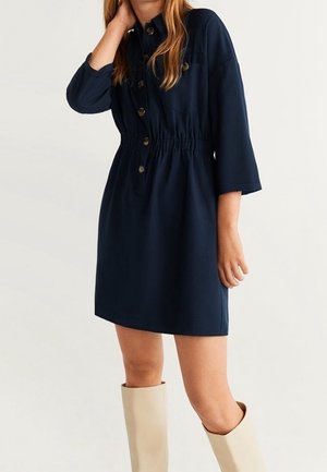 Vrouw draagt een donkerblauwe jurk met knopen en driekwart mouwen en crèmekleurige kniehoge laarzen tegen een effen lichte achtergrond.