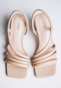 Paar beige flache Sandalen mit quadratischen Zehen und gepolsterten Schlaufenriemen auf weißem Hintergrund.
