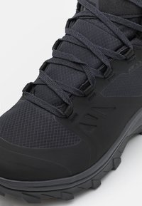 Close-up of a black hiking boot showing laces, textured fabric upper, reinforced toe, and rugged sole tread.
