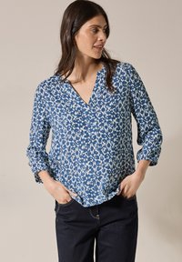 Blå och vit mönstrad blus med V-ringning, långa ärmar uppvikta till armbågarna och en lös passform. Tyget verkar lätt och texturerat.