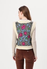 Schwarze Weste mit buntem floralem Stickmuster, cremefarbenem Fleecebesatz und tailliertem Schnitt. Diniert über einem gerippten beigen Pullover, mit braunen Hosen.