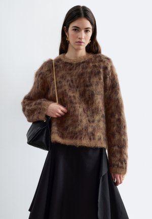 Femme portant un pull marron duveteux, une jupe noire asymétrique, des bottines noires à talons, et portant un sac à main noir avec une bandoulière chaîne dorée.