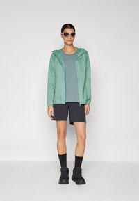 Lightweight, mint green rain jacket with hood, gray athletic top, black shorts, black boots, and sunglasses, showcasing a casual outdoor outfit.