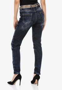 Jean en denim délavé foncé avec une coupe slim, des coutures détaillées, des poches arrière et une ceinture décorative avec des accents de strass. Assorti à des escarpins noirs.