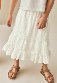 Jupe blanche brodée avec un ourlet volanté et de délicats motifs floraux. Associée à des sandales beige avec des bretelles ajustables et une semelle épaisse.