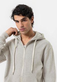 Felpa grigia con zip, tasca frontale, cappuccio con cordino e polsini a costine. Texture liscia e particolare dettaglio della zip.