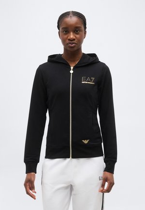 Junge Person trägt einen schwarzen Kapuzenpullover mit Reißverschluss, verziert mit goldenen EA7 Emporio Armani Logos, und weiße Hose mit schwarzen und grünen Streifen.