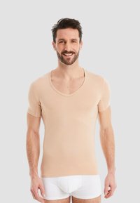 Beige short-sleeve V-neck top made of soft fabric with a fitted design. Model wears white undergarments, standing against a neutral background.