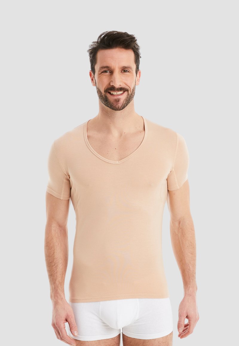 Beige short-sleeve V-neck top made of soft fabric with a fitted design. Model wears white undergarments, standing against a neutral background.