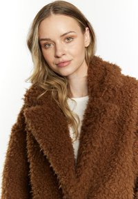 Brun, fluffy frakke med et struktureret stof, bred krave og oversized pasform, båret over en lysfarvet top. Ingen synlige lukninger eller mønstre.