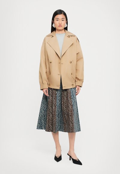 Beige dubbelkleurige jas met oversized revers en elastische manchetten, gecombineerd met een gestructureerde rok met een zigzagpatroon in blauw en bruin.