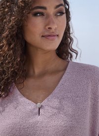 Frau mit lockigem Haar, die ein strukturiertes helllila Oberteil trägt, das mit einem kleinen, runden, mit Schmuck besetzten Knopf geschlossen ist, blickt leicht nach rechts vor klarem Himmel.