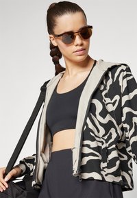 Zebra-mønstret jakke i sort og beige stof med en cropped design, parret med en sort sports-bh. Model iført brune solbriller.