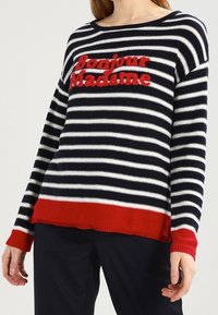 Kobieta ubrana w granatowo-biały sweter w paski z czerwonymi mankietami i dolną krawędzią, z czerwonym haftowanym napisem „Bonjour Madame” na piersi.