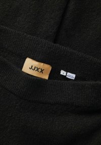 Zwarte gebreide trui met een omgeslagen halslijn. Labels bevatten een gouden tag met "JJXX" en een witte tag met de maat "S" en "Made in Bangladesh."