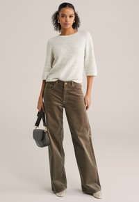 Pantaloni largi din catifea maro, asortați cu un pulover tricotat gri deschis, cu textură. Modelul ține o geantă mică gri cu o curea neagră.