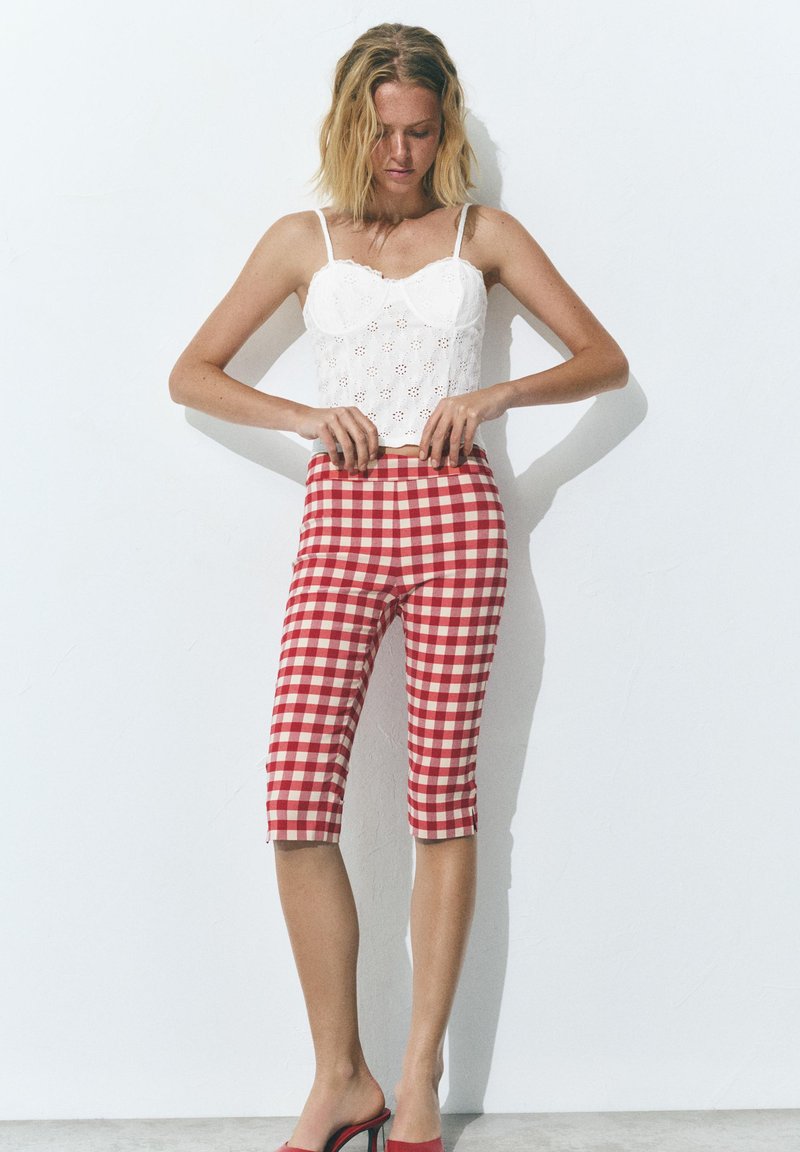 Femme aux cheveux blonds ajustant un pantalon court à carreaux rouges et blancs, portant un haut blanc sans manches et des sandales à talons rouges contre un mur blanc.