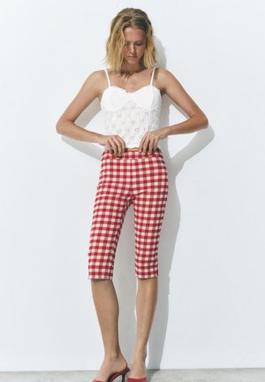 Femme aux cheveux blonds ajustant un pantalon court à carreaux rouges et blancs, portant un haut blanc sans manches et des sandales à talons rouges contre un mur blanc.