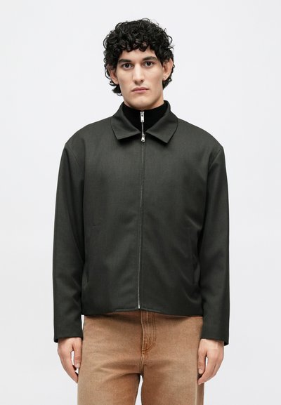 Tmavosivá bunda na zips so štruktúrovaným golierom, dlhými rukávmi a dvoma bočnými vreckami, doplnená o svetlohnedé nohavice. Hladká textúra látky.