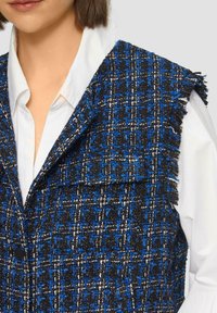Blaues und schwarzes Tweed-Westen mit fransigem Kragen, im karierten Muster und großen vorderen Klappentaschen, über einem weißen Hemd getragen.