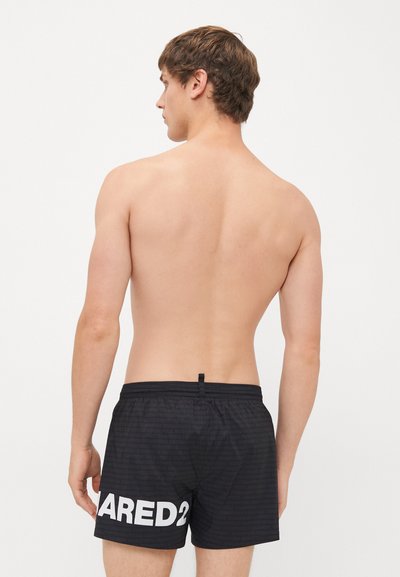 Junger Mann von hinten zu sehen, der schwarze Badehosen mit großem weißen "DSQUARED2"-Schriftzug auf dem linken Bein vor einem schlichten weißen Hintergrund trägt.