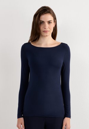 Kobieta stojąca przy białej ścianie, ubrana w dopasowaną granatową bluzkę z długim rękawem i szerokim okrągłym dekoltem, zwrócona twarzą do przodu.