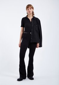 Camisa negra de manga corta con canalé y cuello abotonado, combinada con jeans negros acampanados y una larga chaqueta de mezclilla negra llevada sobre un hombro.