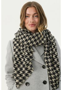 Czarny i kremowy szalik w desenie houndstooth wykonany z miękkiego materiału, z frędzlami na krawędziach. Szalik jest owinięty wokół szarego płaszcza.