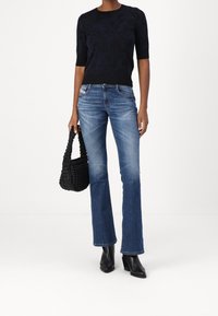 Svart kortärmad texturerad tröja, blå utställda jeans och svart vävd handväska. Modellen bär svarta klackstövlar och står mot en vit bakgrund.