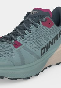 Chaussure de running bleu clair et gris foncé avec des touches roses, des lacets gris, une tige en mesh et le logo "DYNAFIT" sur le côté.