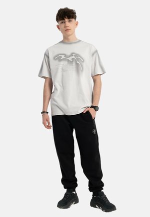 Jasnoszara koszulka z krótkim rękawem z abstrakcyjnym czarnym wzorem graficznym, połączona z czarnymi spodniami dresowymi. Czernie sneakersy o fakturowanej podeszwie dopełniają całość.